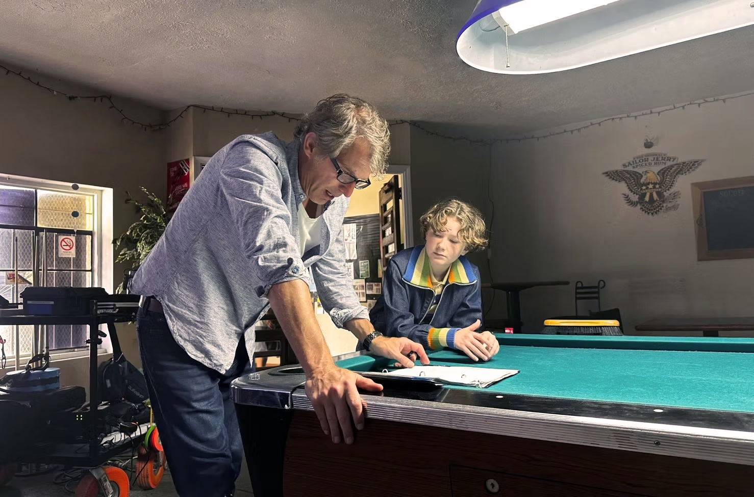 David leans over a pool table, talking through a scene with the young actor on the set of The Alibi