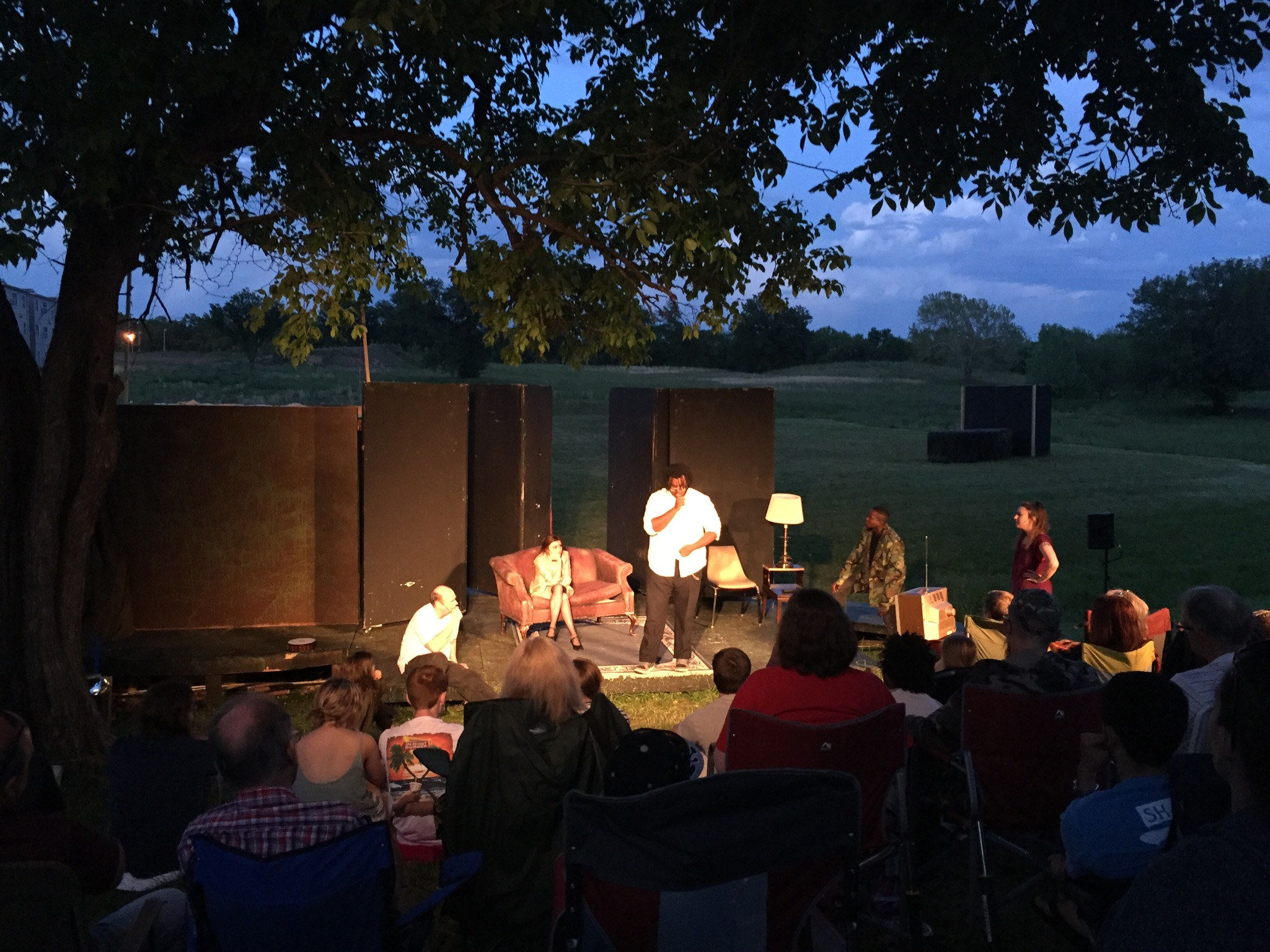 Act two: a lit living-room set under a tree at dusk, the audience on the grass
