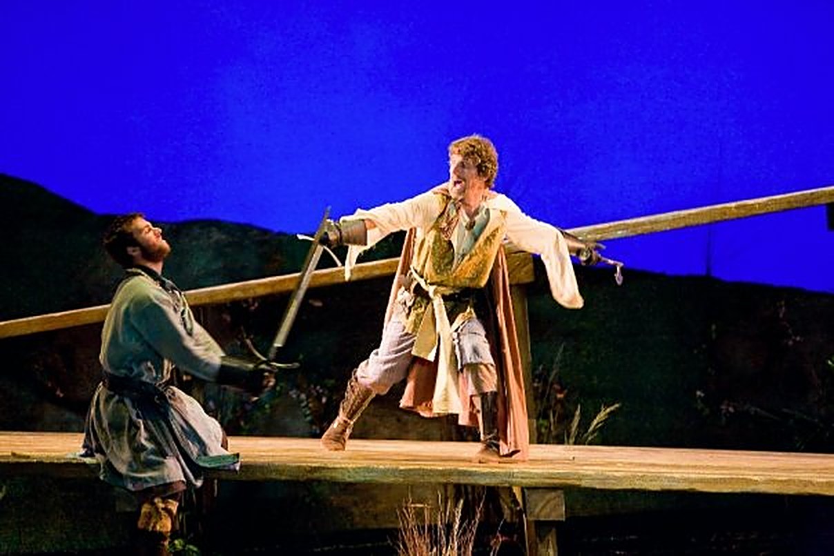 Two actors in a sword fight on a raised wooden platform against a deep blue sky