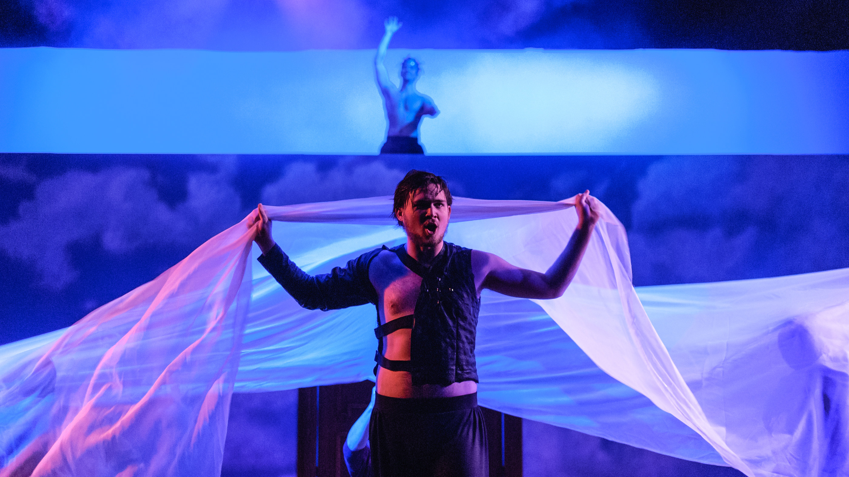 An actor in Metamorphoses holds a length of sheer fabric overhead under blue light